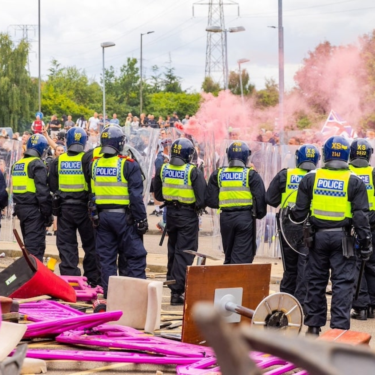 A line of uniformed riot police officers stand in front of damaged furniture and plastic barriers outside a hotel. In the distance are hundreds of protesters, pink smoke and flags